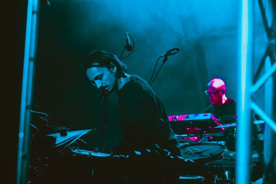 Ein Musiker steht auf einer dunkel beleuchteten Bühne am Mikrofon, umgeben von Schlagzeug und technischem Equipment. Im Hintergrund spielt ein weiterer Musiker an einem Keyboard, während blaues und pinkes Licht die Szene prägen.