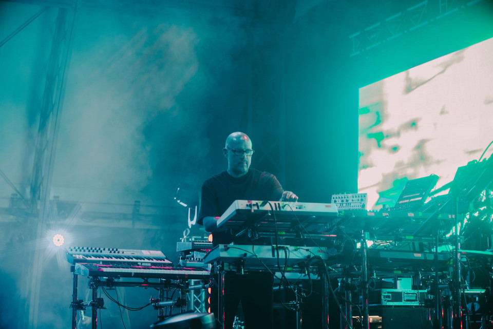 Ein Musiker steht auf einer nebligen Bühne und bedient mehrere Keyboards und Synthesizer. Türkisfarbenes Licht, Bühnenrauch und eine große helle Leinwand im Hintergrund erzeugen eine atmosphärische Konzertstimmung.