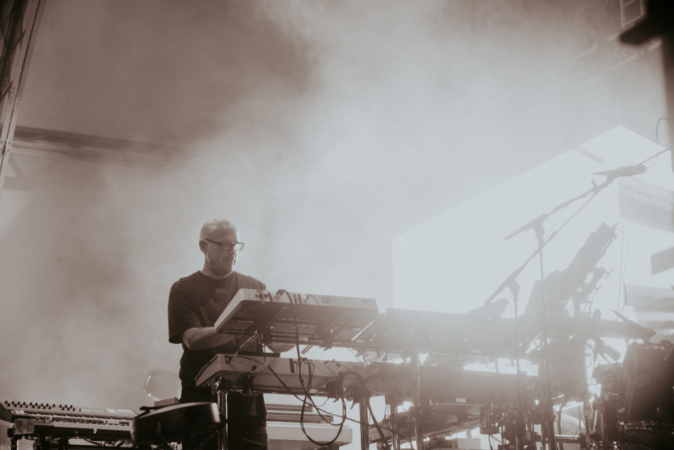 Ein Musiker steht auf einer nebligen Bühne und bedient mehrere Synthesizer und Keyboards. Helles Gegenlicht und Mikrofonständer im Hintergrund erzeugen eine atmosphärische Konzertszene.