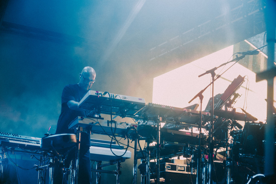 Ein Musiker steht auf einer dunkel beleuchteten Bühne und spielt an mehreren Keyboards und Synthesizern. Blaues Licht, Nebel, Kabel und Mikrofonständer erzeugen eine atmosphärische Konzertszene.
