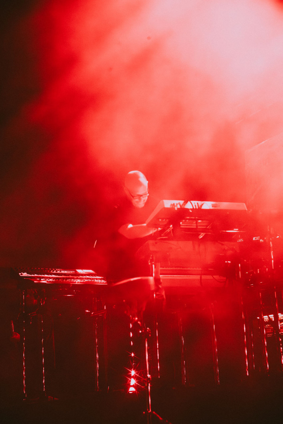 Ein Musiker steht auf einer dunklen Bühne hinter elektronischen Instrumenten und einem Laptop. Rotes Licht und dichter Nebel tauchen die Szene in eine dramatische, fast abstrakte Atmosphäre.