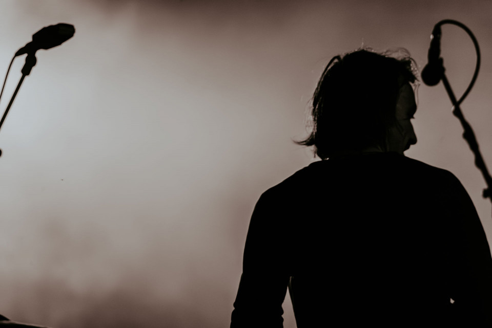 Eine Person ist als dunkle Silhouette vor einem hellen, verschwommenen Hintergrund zu sehen. Links und rechts stehen Mikrofone auf Stativen, was auf eine Bühnen- oder Aufnahmesituation hindeutet.