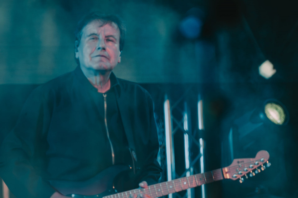 Ein älterer Musiker steht auf einer dunkel beleuchteten Bühne und hält eine elektrische Gitarre. Blaugrünes Licht, Scheinwerfer und ein unscharfer Hintergrund erzeugen eine atmosphärische Konzertstimmung.