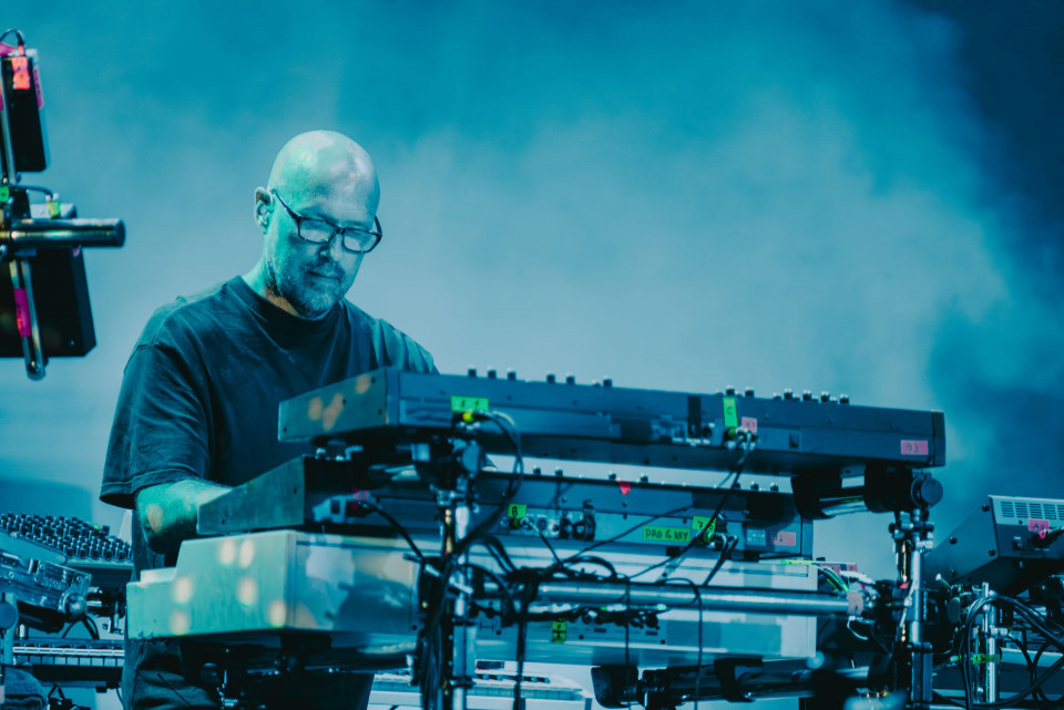 Ein kahlköpfiger Musiker mit Brille steht auf einer blau beleuchteten Bühne und bedient mehrere elektronische Keyboards und Geräte. Um ihn herum sind Kabel, Mischpulte und technisches Equipment zu sehen, während im Hintergrund Nebel oder Projektionen eine atmosphärische Konzertstimmung erzeugen.