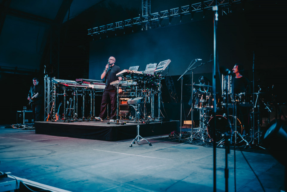 Eine Band steht auf einer dunkel beleuchteten Bühne mit Synthesizern, Gitarren und einem Schlagzeug. Der Sänger hält ein Mikrofon in der Hand, während die Musiker von blauem Bühnenlicht umgeben sind.