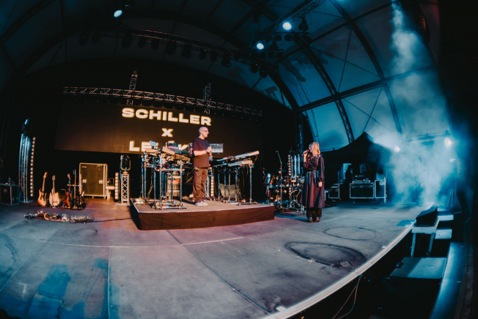 Auf einer großen Konzertbühne stehen zwei Musiker zwischen Keyboards, Schlagzeug und Gitarren, während im Hintergrund „SCHILLER X …“ auf einer LED-Wand leuchtet. Blaues Bühnenlicht und Nebel erzeugen eine atmosphärische, futuristische Stimmung.