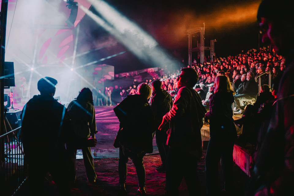 Eine große Menschenmenge sitzt und steht bei einem nächtlichen Konzert vor einer hell beleuchteten Bühne. Bunte Scheinwerfer, Nebel und kräftige Lichtstrahlen tauchen die Szene in eine lebendige Atmosphäre.