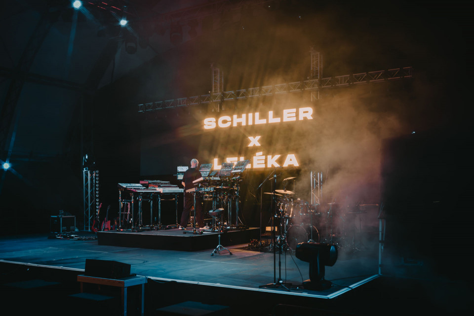 Auf einer dunklen Konzertbühne steht ein Musiker zwischen Synthesizern und elektronischem Equipment, während Nebel und blau-oranges Licht die Szene füllen. Im Hintergrund leuchtet groß der Schriftzug „SCHILLER x LIZÉKA“, daneben sind Schlagzeug, Mikrofone und weitere Bühneninstrumente zu sehen.