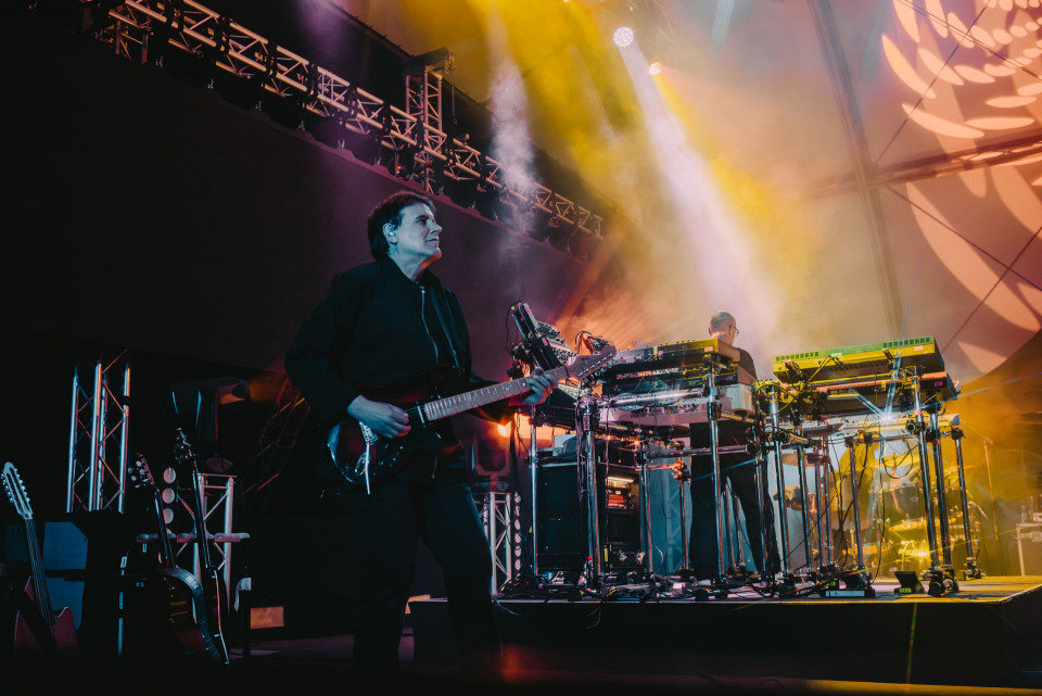 Ein Musiker spielt auf einer Bühne E-Gitarre, während im Hintergrund ein weiterer Künstler an mehreren Keyboards und elektronischen Geräten steht. Bunte Scheinwerfer, Nebel und große Lichtprojektionen erzeugen eine dramatische Konzertatmosphäre.