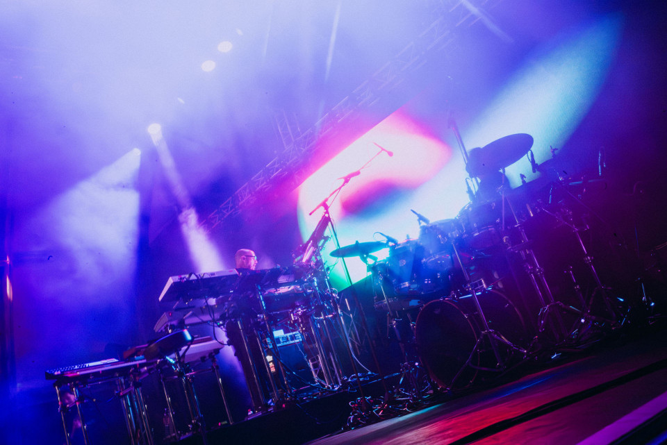 Auf einer Konzertbühne stehen ein Schlagzeug, Keyboards und Mikrofone im intensiven violett-blauen Licht. Bunte Scheinwerfer und Nebel erzeugen eine dynamische, atmosphärische Live-Performance-Stimmung.