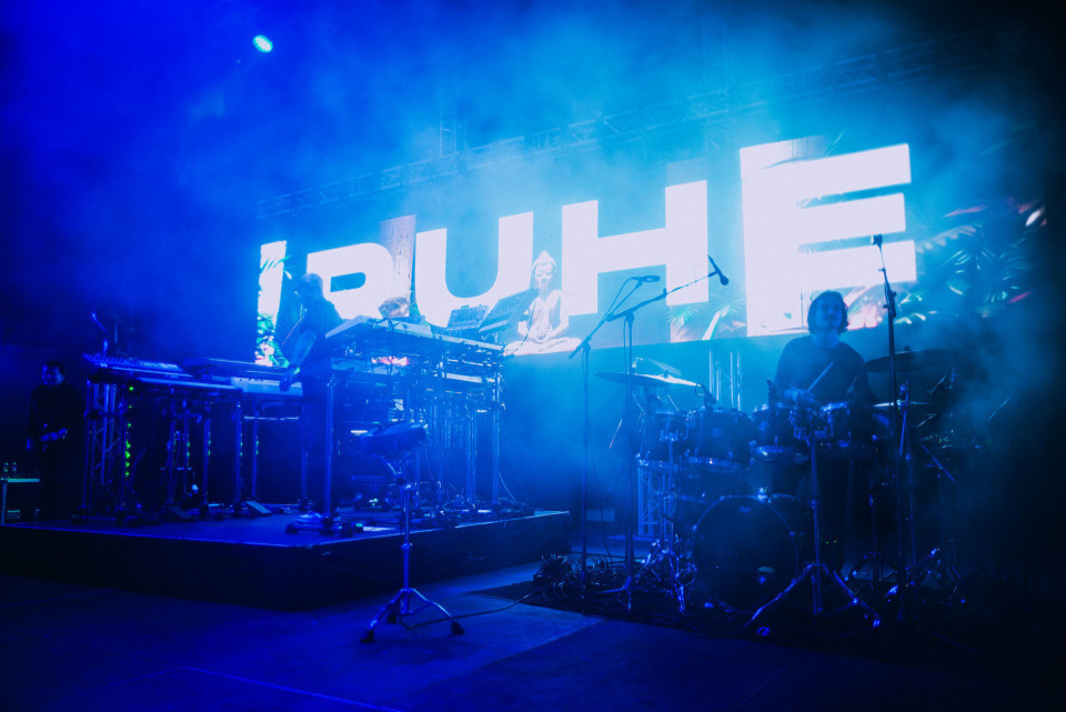 Eine Band spielt auf einer dunkel beleuchteten Bühne, die in intensives blaues Licht und Nebel getaucht ist. Im Hintergrund leuchtet eine große LED-Wand mit riesigen weißen Buchstaben, während Schlagzeug, Mikrofone und elektronische Instrumente zu sehen sind.