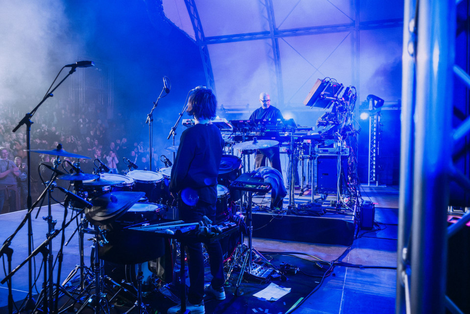 Auf einer blau beleuchteten Bühne stehen Musiker zwischen Schlagzeug, Mikrofonen, Synthesizern und vielen Kabeln. Links ist ein großes Publikum zu sehen, während Nebel und Scheinwerfer die Konzertatmosphäre verstärken.