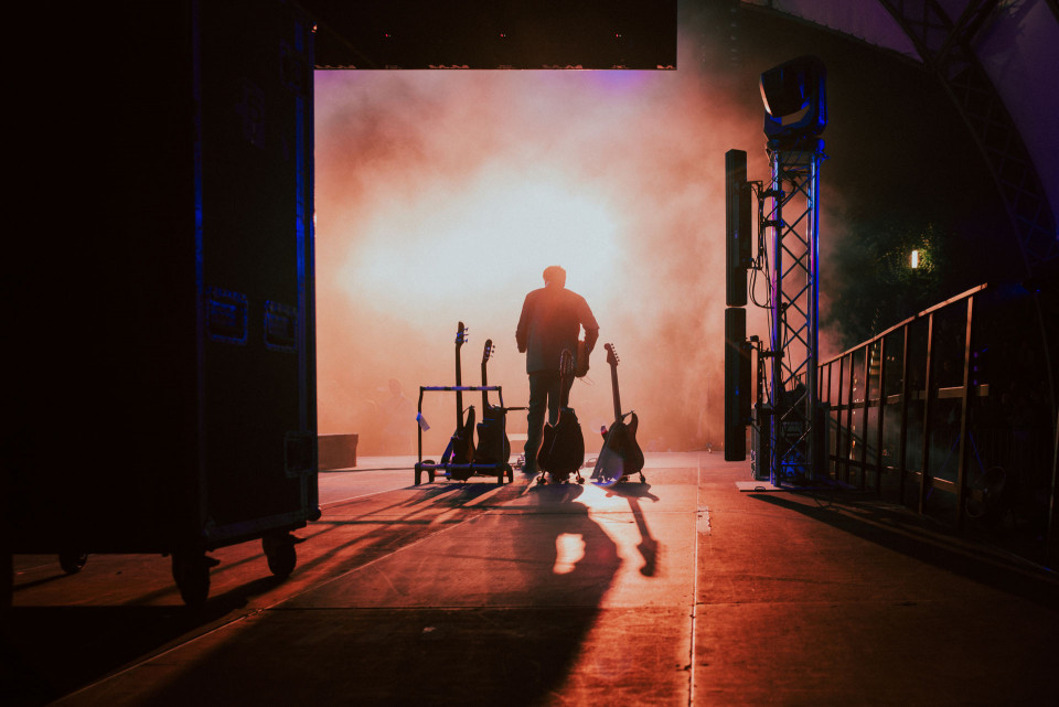 Ein Musiker steht als Silhouette auf einer Bühne, umgeben von mehreren Gitarren und dichtem Bühnennebel. Warmes Gegenlicht und blaue Akzente beleuchten die Konzertszene hinter den Kulissen.
