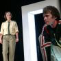 Ein Mann in einem futuristischen Anzug steht vor einer großen, weiß erleuchteten Tür, während eine Frau in Uniform ihn aus der Ferne betrachtet. Das Bild scheint Teil einer Theaterproduktion oder einer Inszenierung zu sein, die in einem dunklen Hintergrund spielt.