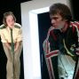 Zwei Schauspieler stehen vor einer großen, weißen Tür im Hintergrund, einer in Uniform eines Parkwächters und der andere in einer auffälligen Jacke. Die beiden blicken in unterschiedliche Richtungen und scheinen sich zu unterhalten oder zu beobachten.