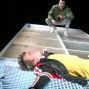 Ein Mann steht auf einer weißen, quadratischen Plattform und deutet auf einen anderen Mann, der auf dem Boden liegt und dabei eine rote Jacke trägt. Im Hintergrund sind mehrere Lichtquellen sichtbar, die das Bühnenbild beleuchten.