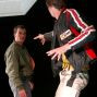 Ein Mann in einer grünen Jacke und ein anderer Mann in einer gelb-schwarzen Motorradjacke stehen vor einem dunklen Hintergrund und scheinen miteinander zu diskutieren. Beide Männer scheinen in einer theatralischen oder improvisierten Szene zu sein.