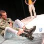 Ein junger Mann sitzt auf einer weißen, mit einem Fischgratmuster versehenen Fläche und trägt dabei schwarze Stiefel. Neben ihm steht ein weiterer Mann in einem roten und weißen Jackett und Jeans, der auf einer ähnlichen Fläche steht.