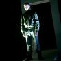 Ein junger Mann in einem grünen Jackett und Jeans steht vor einem dunklen, rechteckigen Durchgang. Die Szene ist von einem starken, grünen Licht beleuchtet, das ihn hervorhebt und den Hintergrund in Schatten taucht.