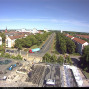 Die Aufnahme zeigt eine breite, von Bäumen gesäumte Straße in einer Stadt mit Wohnhäusern, Kirche und mehreren Autos unter klarem blauem Himmel. Im Vordergrund befindet sich ein Baustellenbereich auf einem Dach oder erhöhten Platz mit Gerüsten, Baumaterialien und arbeitenden Personen.