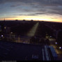 Das Foto zeigt eine Stadtlandschaft in der Dämmerung mit einer langen, beleuchteten Straße, die zum Horizont führt. Am Himmel sind dunkle Wolken und ein heller Streifen des Sonnenuntergangs zu sehen, während Gebäude und Bäume bereits im Schatten liegen.