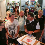 Auf einem Messegelände stehen viele Menschen vor einem Stand, auf dem ein Mann einen Besucher berät. Im Hintergrund sind weitere Besucher und ein Fahrzeug zu sehen, während einige Personen im Gespräch stehen.