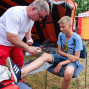 Ein älterer Mann versorgt einen Jungen, der sich an der Wade verletzt hat, während sich im Hintergrund ein Campingzelt und Fahrräder befinden. Beide Personen sitzen auf Stühlen, während der Mann ein Pflaster anbringt.