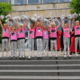 Eine Gruppe junger Cheerleaderinnen in pinken Shirts mit silberfarbenen Akzenten und silbernen Leggings stehen auf einer Treppe vor einem Gebäude. Sie halten ihre Arme in der Luft und scheinen bei einer Aufführung oder einem Event zu erscheinen.