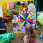 Auf einem Festgelände vor einem Hintergrund aus Bäumen steht ein Mann und ein kleines Kind vor einem großen Spielstand mit einer Wendelrad und einigen Spielen. Es werden zudem Luftballons und verschiedene Aufsteller mit Kinderbildern und Logos zu sehen sind.