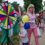 Auf einem Marktplatz steht eine Frau mit einem Ballon und einem kleinen Kind vor einem großen Tamburin, auf dem die Zahlen 1 bis 16 angeordnet sind. Im Hintergrund befinden sich weitere Besucher und ein Fahrrad.