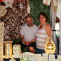 Ein Paar lächelt fröhlich vor einem Stand, der mit Holz, Kerzen und Schmuck verziert ist. Der Stand ist mit einem Wand aus Holzscheiben und einer grünen Plane im Hintergrund ausgestattet.