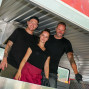 Drei Mitarbeiter, ein Mann und zwei Frauen, stehen lächelnd im Innenraum eines roten Food Trucks. Sie tragen schwarze T-Shirts und ein Schürze, und der Truck scheint ein mobiler Imbissstand zu sein.