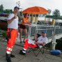 Auf einem Hafenufer stehen ein Mann in orangefarbenem Warnschutzanzug und zwei Frauen, die ebenfalls Warnbekleidung tragen, lächelnd vor einem Boot. Ein Mann sitzt in einem Rollstuhl auf einer Bank und ein weiteres Boot ist im Hintergrund zu sehen.