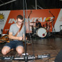 Ein Musiker sitzt auf einer Bühne und spielt mit einer Gitarre, während im Hintergrund eine Schlagzeuggruppe steht. Vor ihnen ist eine große orangefarbene Banner mit Werbeaufdrucken und Logos zu sehen.