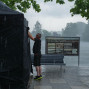 Ein Mann deckt einen Bollwerk im Regen ab, während draußen ein regnerischer Tag herrscht. Im Hintergrund ist ein See und eine historische Stadt zu sehen, die vom Nebel umhüllt ist.