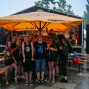 Eine Gruppe von Menschen unter einem Biergartenpavillon lacht und posiert im Regen. Sie sind in T-Shirts und Shorts gekleidet und haben Getränke und Becher auf den Tischen vor sich.