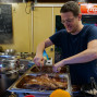 Ein junger Mann mit Brille lächelt und betrigt mit Gabel und Messer Fleisch in einer großen Dampfschüssel. Hinter ihm befindet sich eine Theke mit Speisekarte und einer Kaffeemaschine, die ein Restaurant darstellt.