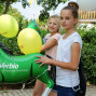 Zwei junge Mädchen posieren vor einem großen, grünen Heliumpferd mit dem Logo "Verbio". Sie halten sich gegenseitig über die Schulter, während im Hintergrund ein Gebäude und Ballons zu sehen sind.