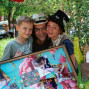 Eine Familie mit einem Jungen und einer Frau lächelt vor einem dekorativen Bild mit Fotos und Blumen. Ein älterer Mann trägt einen Hut und hält ebenfalls das Bild in der Hand und blickt in die Kamera.