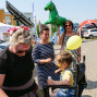 Auf einem belebten Marktplatz stehen eine Gruppe von Menschen vor einem großen, aufblasbaren Pferd, während ein kleines Kind mit einem Ballon spielt. In der Mitte der Gruppe stehen Frauen, die auf ein Foto lächeln und das Kind beobachten.