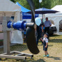 Auf einem Festplatz steht ein großer Propeller auf einem Stahlgestell, umgeben von Informationsplakaten und einem Zelt. Ein kleiner Junge schaut fasziniert auf den Propeller, während ein Mann neben ihm steht und eine Limousine betrachtet.