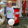 Ein kleiner Junge steht vor einem großen, aus Rohren gebauten Roboter und hält neben sich eine weiße Kugel. Im Hintergrund ist ein Getränkestand und weitere Personen zu sehen, alles auf einem Festival oder einer Veranstaltung.