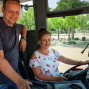 Ein Mann und eine Frau stehen auf einem Bus und lächeln in die Kamera. Die Frau sitzt im Fahrersitz und bedient das Lenkrad, während der Mann an ihrem Rücken lehnt.