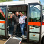 Zwei Personen, ein Mann und eine Frau, posieren fröhlich vor einem weißen Bus mit dem Schriftzug "TheaterBus". Der Bus ist mit Firmenlogos und Werbeaufdrucken versehen und dient als mobile Büros bzw. Wartebereiche.
