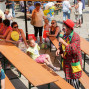 Auf einem Marktplatz unterhalten sich viele Menschen, während ein Clown mit einer kleinen, glücklichen Mädchen interagiert. Ein großer Smurfs-Luftballon steht neben einem Tisch mit Eis und einer Straßenbank mit einer Gruppe von Kindern.