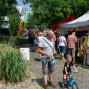 Auf einem belebten Markt, der unter einem Zelt steht, unterhalten sich ein Mann mit seinem Sohn, der auf einem kleinen Fahrrad fährt, während andere Besucher die Stände erkunden. Im Hintergrund sind mehrere Marktstände mit Waren und eine Gebäudefront zu sehen.