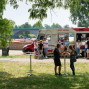 Auf einer Wiese vor einem Brückenparkplatz steht ein Foodtruck mit mehreren Kunden, die sich zum Essen versammeln. Hinter dem Truck stehen mehrere Personen um Tische, während weitere Kunden im Hintergrund stehen und warten.