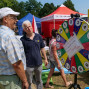 Zwei ältere Männer unterhalten sich vor einem farbenfrohen Gewinnrad, das von einem Rotary-Club-Event zu sehen ist. Im Hintergrund sind weitere Personen und Kinder auf einem Freibereich zu erkennen.