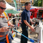 Zwei Männer, einer mit orangefarbenem Sicherheitsgurt, betrachten eine Feuerwagen-Demonstration auf einem öffentlichen Platz. Im Hintergrund stehen Menschen und Kinder, während ein Sonnenschirm für Schatten sorgt.