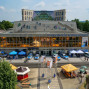 Auf dem Foto ist ein Mehrfachparkplatz mit zahlreichen Polizeiautos und einem öffentlichen Biergarten vor dem Theaterklause in Luckenau zu sehen. Viele Menschen versammeln sich auf dem Parkplatz und im Biergarten, während im Hintergrund ein moderner Gebäudekomplex mit einer Treppe und Fenstern zu erkennen ist.